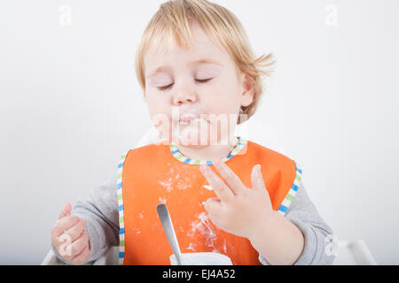 blonde caucasian baby seventeen month age orange bib grey sweater eating meal with her hand in white high chair Stock Photo