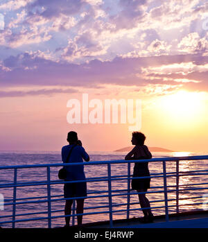 Silhouettes of a man and a woman taking pictures and looking at the sunset on  sea and island from ferry deck Stock Photo