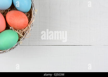 High angle shot of a basket of Easter Eggs in the corner of the frame. The basket is on a rustic white wood kitchen table. Stock Photo