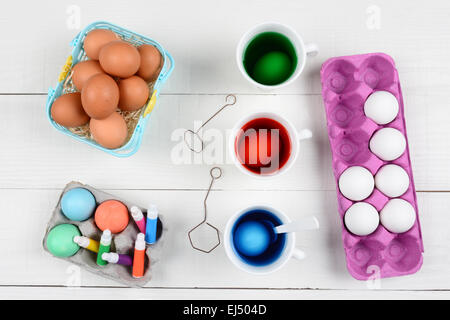 High angle still life of dying eggs for Easter. Three cups with dye, a basket of brown eggs and cartons of undyed and dyed eggs Stock Photo