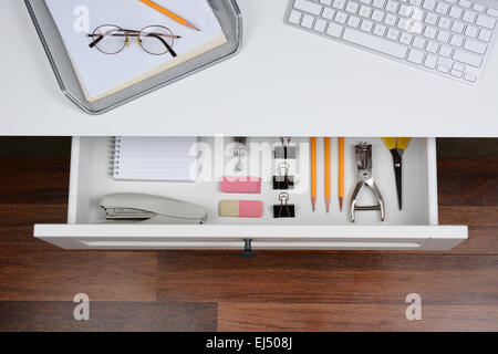 High angle shot of an open desk drawer showing the items inside. The top of the desk has a computer keyboard and wire in-box wit Stock Photo