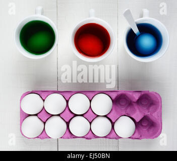 High angle still life of eggs being dyed for Easter. Three cups with dye and an egg in each, a pink carton of un-dyed eggs on a Stock Photo