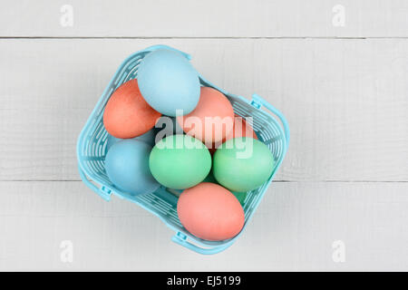 Pastel Easter eggs in a plastic basket on a white rustic wood kitchen table. High angle view with copy space. Stock Photo