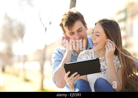 Couple sharing music and singing with a tablet sitting in a bench in an urban park Stock Photo