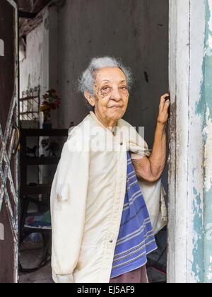 Afro-Cuban female senior citizen with gray hair stands in the doorway of her house looking out with one arm up against the door. Stock Photo