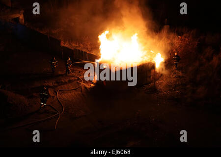 A squad of four firefighters dousing a burning shed and wooden fence with fire hose. The fire takes place in woodland at night a Stock Photo