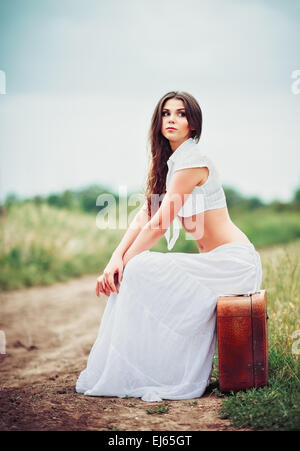 Beautiful young woman sits on a suitcase near field road Stock Photo