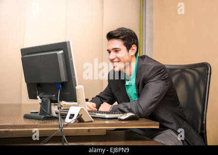 Successful handsome young businessman sitting at his desk in the office smiling and looking at the computer screen Stock Photo