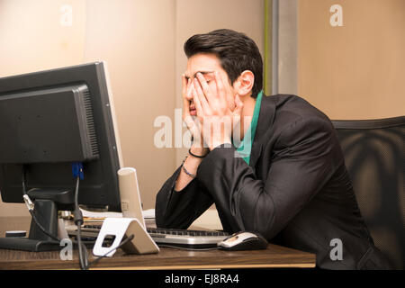 Tired bored young businessman sitting at his desk in front of his computer with his chin resting on his hands and eyes closed