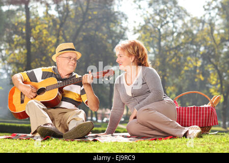 Senior man playing guitar to his wife on a picnic, seated on a blanket. The shot is in a city park during early autumn Stock Photo