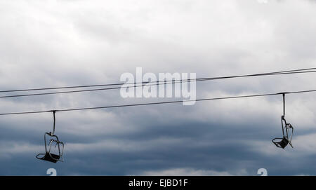 Two empty ski lift chairs hanging on steel cable against overcast sky. Stock Photo