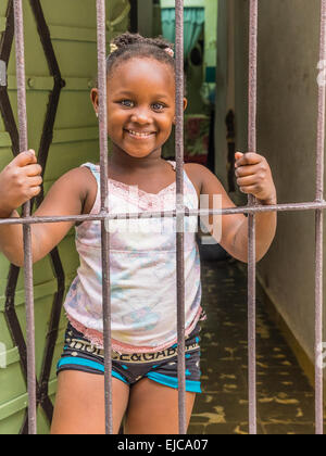 A young Afro-Cuban girl holds the bars of the security door to her house with both hands and looks out to the street and smiles. Stock Photo