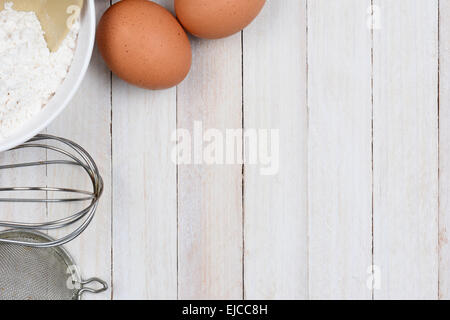 High angle shot of a baking still life, The objects, flour bowl, whisk, strainer and eggs are in the upper left corner, leaving Stock Photo