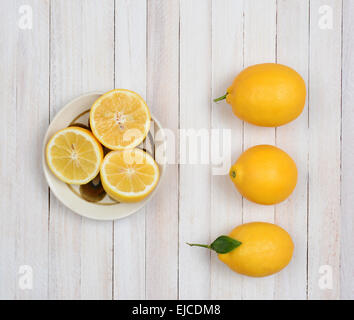 High angle still life of three whole lemons and three lemon halves in a bowl on a rustic white wood kitchen table. Stock Photo