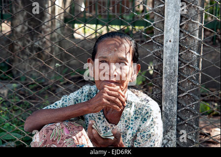 An old Burmese lady begging sitting on the pavement against a wire fence on a Rangoon street Myanmar Burma Stock Photo