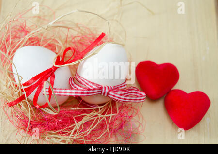 Who white eggs tied with red ribbons lying in a nest. Two small heart shaped pillows on a wooden table. Stock Photo