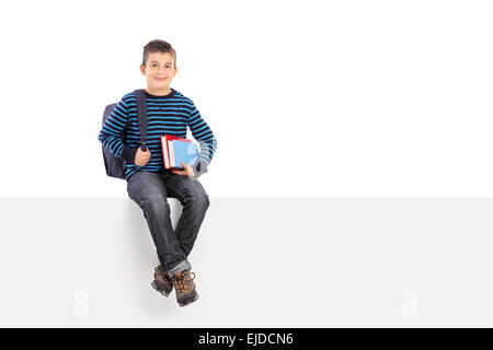 Schoolboy holding a couple of books and carrying a backpack seated on a blank signboard Stock Photo