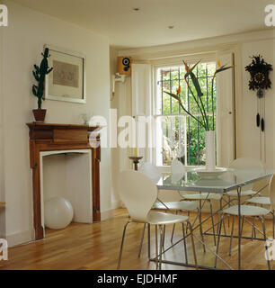 White Arne Jacobsen style chairs at glass topped table in modern dining room with simple pine mantelpiece and old cuckoo clock Stock Photo