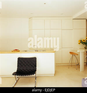 Black woven leather and chrome chair in modern white minimalist kitchen with wall of streamlined storage cupboards Stock Photo