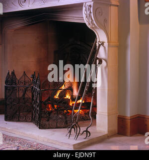Close-up of fireplace with lighted fire and wrought iron fireguard and fire tongs Stock Photo
