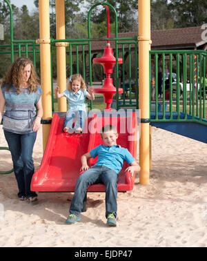 Young boy and girl go down slide on park playground while Mom looks on Stock Photo