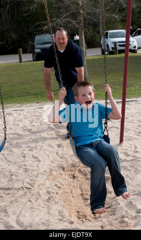 Dad pushes eight year old boy on swing at park playground Stock Photo