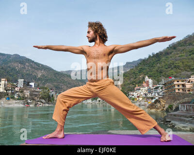 A man practicing yoga warrior pose by the Ganges river in Rishikesh India Stock Photo
