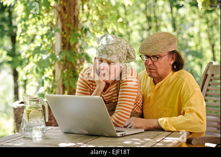 Senior couple outdoors with a laptop, They're looking at the computer. There's a sunny background of trees and bushes Stock Photo
