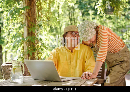 Senior couple outdoors with a laptop, They're laughing. There's a sunny background of trees and bushes Stock Photo