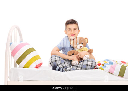 Studio shot of a happy little boy in pajamas, sitting in bed and hugging a teddy bear isolated on white background Stock Photo