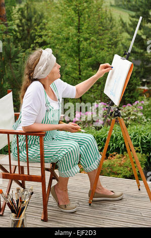 Senior woman painting outdoors. She sits in a canvas chair in front of the easel. The day is overcast giving a soft light Stock Photo