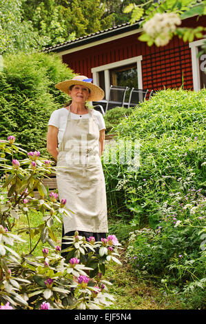 Senior woman standing in her garden, She is surrounded by bushes. There's a red house partly seen in the background. Stock Photo