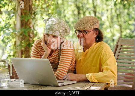 Senior couple outdoors with a laptop, They're looking at the computer. There's a sunny background of trees and bushes Stock Photo
