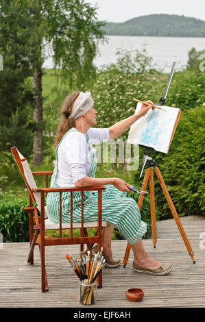 Senior woman painting outdoors. She sits in a canvas chair in front of the easel. The day is overcast giving a soft light Stock Photo