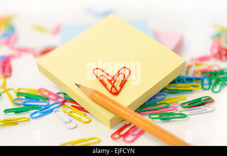 Small note pad and a pencil on a background of multi-colored paper clips on white. Stock Photo