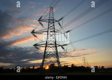 Two forms of energy - setting sun behind electricity pylon and power cables in landscape at sunset in England, UK Stock Photo