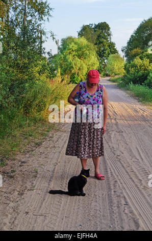 Rural landscape - woman and black cat on sandy road in village. Stock Photo