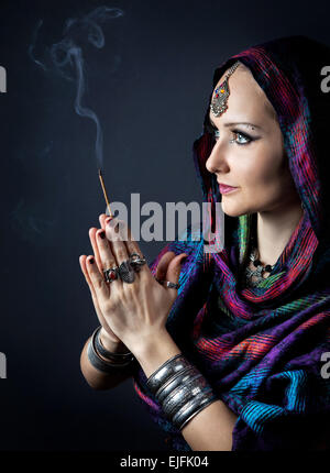 Woman wrapped in scarf holding incense stick in hands with Namaste gesture at dark background Stock Photo