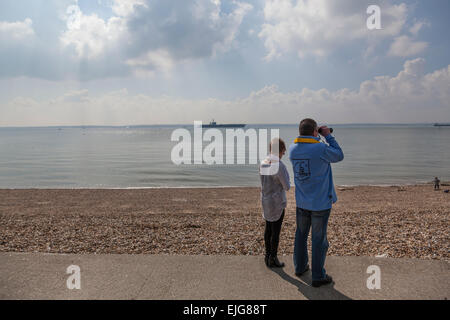 Stokes Bay, Solent, UK. 25th March, 2015. A couple survey the Solent with binoculars as the Nimitz class aircraft carrier USS Theodore Roosevelt lies at anchor in the distance, 25th March 2015. Credit:  Anthony Hatley/Alamy Live News Stock Photo