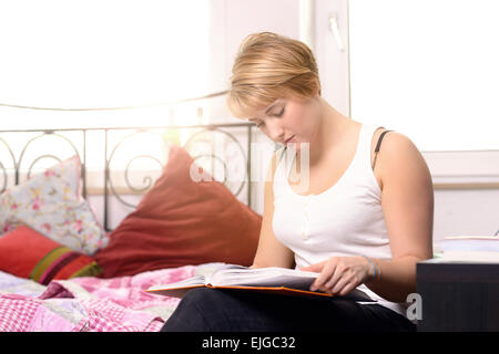 Pretty young female student reading in a textbook while sitting in her bedroom, the sun shining through the window Stock Photo