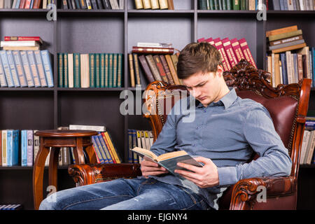Handsome young man sitting reading in a brown leather armchair the library concentrating on his book Stock Photo