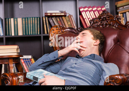 Sleepy young man yawning with his hand to his mouth as he relaxes in a comfortable leather armchair library with his book Stock Photo