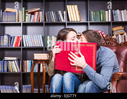 Romantic young couple sneaking a kiss as they sit reading in a library holding up a large red book to partially conceal their fa Stock Photo