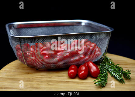 Stainless steel basket filled with plum tomatoes with rosemary sprigs on a bamboo cutting board isolated on black Stock Photo