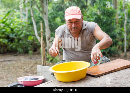 An older man wearing a pink cap and a gray sleeveless shirt is seated outdoors, focused on preparing food. He is using a small knife in one hand and a pink object in the other, working over a wooden Stock Photo