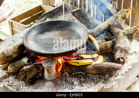 A black, round iron pan is positioned over a small open fire built with logs and kindling, supported by a stone or clay stand. Flames and smoke rise from the fire, which is set within a shallow stone Stock Photo