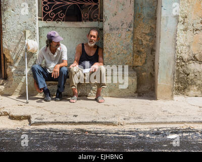 Two Cuban Hispanic male seniors sit on a concrete bench in front of a building in Havana Vieja, Cuba. Stock Photo