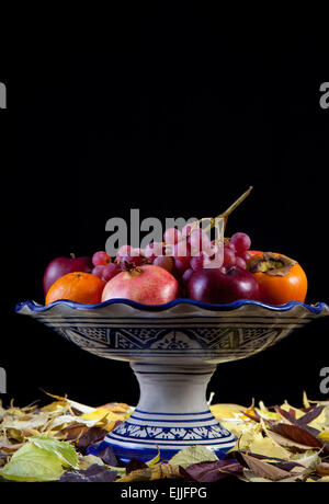 Ceramic fruit bowl with autumn fruits isolated over black background. Surface full of dry leaves Stock Photo