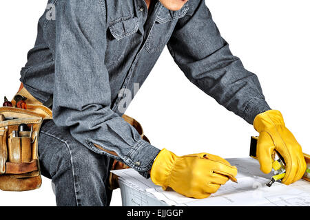 Man in black denim wearing used tool belt filled with carpenter tools reviewing blueprints holding a tape measure and a pencil Stock Photo