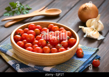 Cherry tomatoes in wooden bowl with garlic, onion, parsley leaves and wooden cutlery in the back Stock Photo